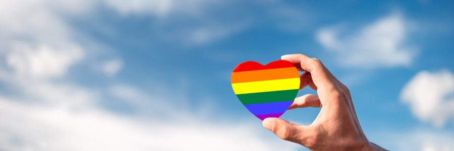 Hand holding painted rainbow heart symbolizing gay and lgbt pride and sky with clouds in the background. Concept of human relationship, support and staying together.